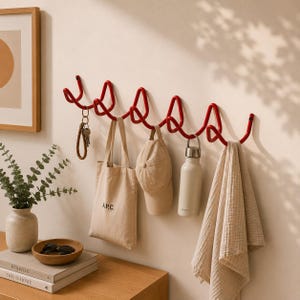 Könnte beinhalten: Eine rote Wandgarderobe aus Metall mit einem durchgehenden, wellenförmigen und geschlungenen Design mit mehreren Haken. Die Garderobe hält eine beige Tragetasche mit dem Text A.P.C., eine beige Baseballkappe, eine silberne Metallflasche und ein beiges Handtuch.