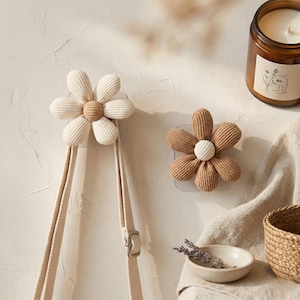 Puede incluir: Dos ganchos de pared con forma de margarita en pana crema y marrón. Un gancho sostiene una correa beige. También se ven una vela encendida en un tarro de cristal y un cuenco pequeño con lavanda.