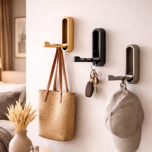 Könnte beinhalten: Drei Wandhaken in Gold, Schwarz und Silber. Eine gewebte Tragetasche hängt am goldenen Haken, Schlüssel am schwarzen Haken und eine Baseballkappe am silbernen Haken. Die Haken sind an einer weißen Wand montiert.