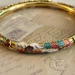 May include: A gold-coloured bangle bracelet with a floral design. The bracelet features colourful enamel flowers in red, teal, and brown against a white background. The bracelet is a decorative jewellery item.