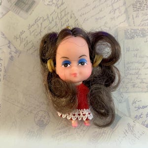 May include: A small doll with brown hair styled in two side ponytails, featuring blue eyeshadow and pink lips. The doll wears a red velvet dress with a white lace trim at the hem. A gold-colored loop is attached to the top of the doll's head.