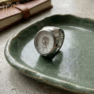 Op de afbeelding: Een zilverkleurige ringhorloge met een witte wijzerplaat met een schedelontwerp. Het horloge heeft een stretchband en toont de cijfers 12, 3, 6 en 9. De ring ligt op een groene, getextureerde plaat.