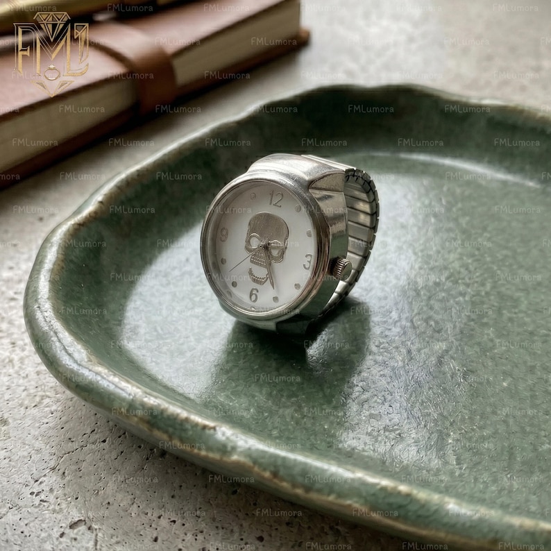 Op de afbeelding: Een zilverkleurige ringhorloge met een witte wijzerplaat met een schedelontwerp. Het horloge heeft een stretchband en toont de cijfers 12, 3, 6 en 9. De ring ligt op een groene, getextureerde plaat.