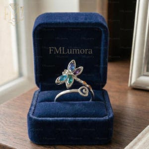 May include: A silver dragonfly ring with blue, green, and purple gemstone accents, presented in a blue velvet ring box. The ring has a delicate design with a curved band.