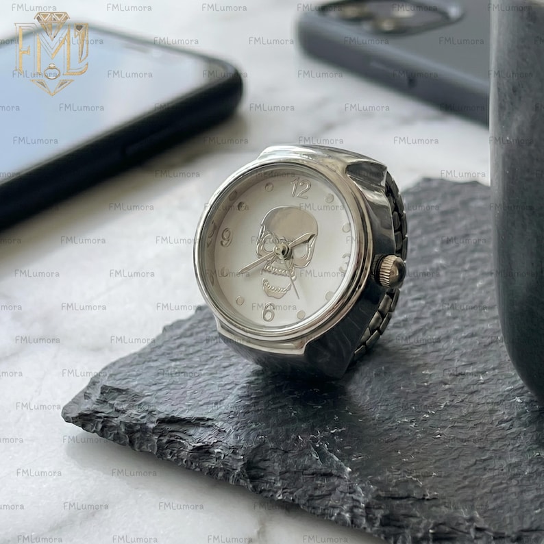 Op de afbeelding: Zilverkleurige ringhorloge met een witte wijzerplaat met een schedelontwerp. Het horloge heeft een ronde wijzerplaat met zwarte wijzers en een kleine kroon. De ring ligt op een zwart oppervlak.