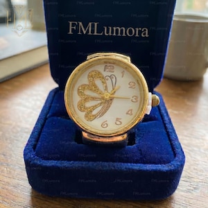 May include: A gold ring watch with a white face and a gold butterfly design. The watch has black numerals and hands. The ring is displayed in a blue velvet box. The brand name "FMLumora" is visible.