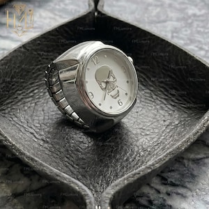 Op de afbeelding: Een zilverkleurige ringhorloge met een witte wijzerplaat met een schedelontwerp. Het horloge heeft zwarte wijzers en de cijfers 12, 3, 6 en 9 zijn zichtbaar. De ring ligt op een donkergrijze leren tray.
