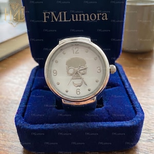 May include: A silver-toned watch with a white face featuring a skull design. The watch has a round face with the numbers 3, 6, 9, and 12. The watch is displayed in a blue velvet box. The brand name "FMLumora" is visible.