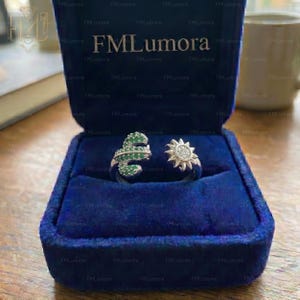 May include: Two silver rings displayed in a blue velvet ring box. One ring is shaped like a snake with green gemstones, and the other is a sunburst design with small, clear stones. The box has the text "FMLumora" on the inside.
