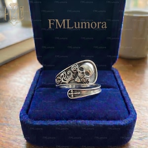 Peut inclure: Bague argentée ornée d'un crâne et de roses, présentée dans un écrin de velours bleu foncé. La bague a un anneau réglable avec un motif de croix. De style gothique ou alternatif.
