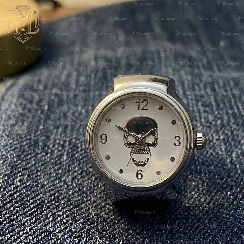 Op de afbeelding: Een zilverkleurige ringhorloge met een witte wijzerplaat met een schedelmotief. De wijzerplaat toont de cijfers 12, 3, 6 en 9. Het horloge ligt op een blauwe denim achtergrond. De diameter van het horloge is ongeveer 2,5 cm.