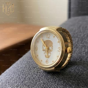 Op de afbeelding: Goudkleurige ringhorloge met een witte wijzerplaat met een schedelontwerp. Het horloge heeft een stretchband en toont de cijfers 12, 3, 6 en 9. Een uniek accessoire.