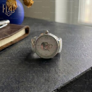 Op de afbeelding: Een zilverkleurige ring die is ontworpen om op een polshorloge te lijken. De ring heeft een ronde wijzerplaat met een gedetailleerd ontwerp en zichtbare wijzers. De band van de ring is breed en solide, met een klein kroontje aan de bovenkant.
