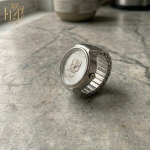 Op de afbeelding: Een zilveren ringhorloge met een schedelontwerp op de wijzerplaat. Het horloge heeft een ronde wijzerplaat met een schedel in het midden en de cijfers 12, 3, 6 en 9. De band is gemaakt van zilvermetaal en is rekbaar.
