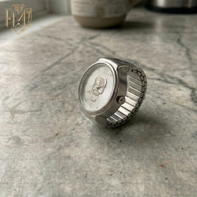 Op de afbeelding: Een zilveren ringhorloge met een schedelontwerp op de wijzerplaat. Het horloge heeft een ronde wijzerplaat met een schedel in het midden en de cijfers 12, 3, 6 en 9. De band is gemaakt van zilvermetaal en is rekbaar.