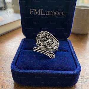 May include: A silver-colored ring with a sunflower design. The ring is in a blue velvet box. The sunflower is in the center of the ring, with leaves and stems wrapping around the band. The ring is adjustable.