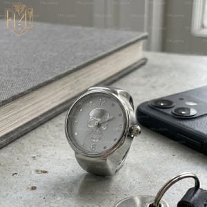 Op de afbeelding: Een zilveren ringhorloge met een witte wijzerplaat met een schedelontwerp. De ronde wijzerplaat toont de cijfers 12, 3, 6 en 9. Het horloge staat tegen een vage achtergrond met een boek en een smartphone.