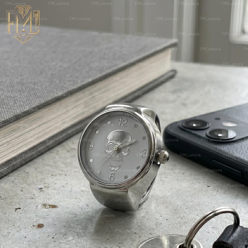 Op de afbeelding: Een zilveren ringhorloge met een witte wijzerplaat met een schedelontwerp. De ronde wijzerplaat toont de cijfers 12, 3, 6 en 9. Het horloge staat tegen een vage achtergrond met een boek en een smartphone.