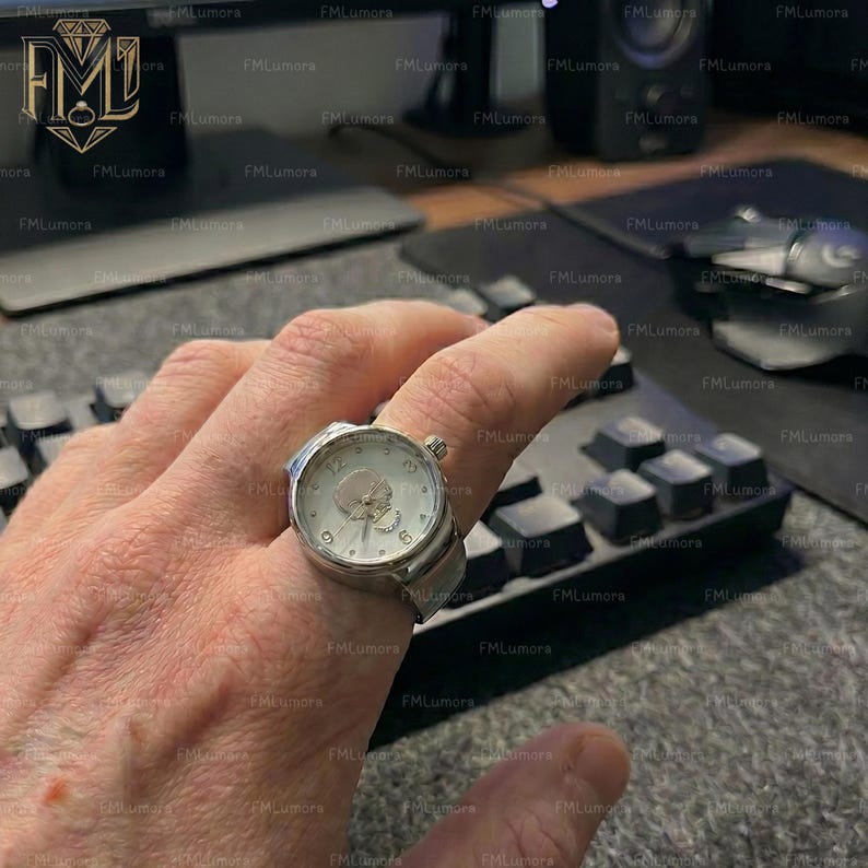 Op de afbeelding: Een zilveren ringhorloge met een witte wijzerplaat en zwarte cijfers. Het horloge heeft een kleine kroon en een band die om de vinger gewikkeld is. Het horloge zit om de vinger van een persoon.