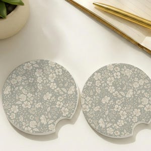 May include: Two circular car cup holder coasters featuring a sage green background with a repeating pattern of small white flowers and leaves. Each coaster has a small semi-circular notch on the edge for easy removal from a vehicle cup holder.