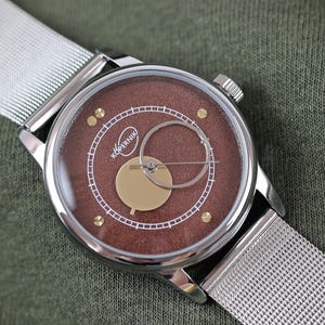 Op de afbeelding: Een zilveren polshorloge met een bruine wijzerplaat en een zilveren mesh band. De wijzerplaat heeft gouden accenten en het woord "KOPERNIK" in het wit. Het horloge ligt op een groene stoffen achtergrond.