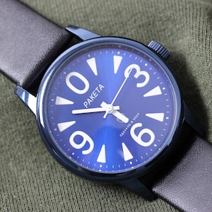 Puede incluir: Un reloj de pulsera con esfera azul con números y agujas blancas, y una correa de cuero negro. La esfera del reloj tiene la palabra "PAKETA" impresa. El reloj está sobre un fondo de tela verde.
