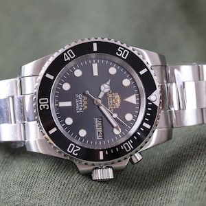 May include: A stainless steel wristwatch with a black dial and a black rotating bezel. The watch features luminous circular hour markers, silver-toned hands, and a day-date window at the three o'clock position. The dial displays the text Orient, Crystal, and 21 Jewels.