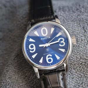 Peut inclure: Montre-bracelet avec un cadran bleu et des chiffres blancs. Le boîtier est argenté et le bracelet en cuir noir. Le mot "PAKETA" est imprimé sur le cadran. Une montre classique.