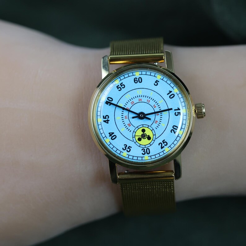 May include: A gold-tone wristwatch with a light blue dial featuring black numerals for minutes, a smaller inner circle with red numerals for hours, and a yellow circular icon at the bottom. The watch has a gold-tone mesh strap and a side crown.