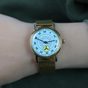 May include: A gold-tone wristwatch with a light blue dial featuring black numerals from 5 to 60, a central 24-hour scale, and a yellow radiation warning symbol at the 6 o'clock position. The watch has thin black hands and a gold-tone mesh metal strap.