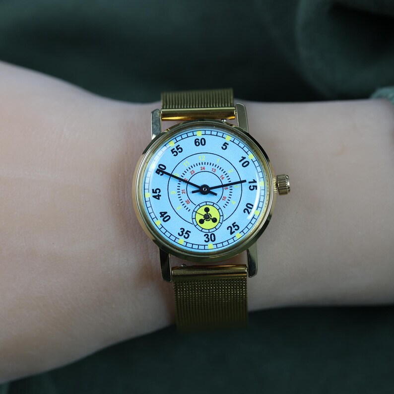 May include: A gold-tone wristwatch with a light blue dial featuring black numerals from 5 to 60, a central 24-hour scale, and a yellow radiation warning symbol at the 6 o'clock position. The watch has thin black hands and a gold-tone mesh metal strap.