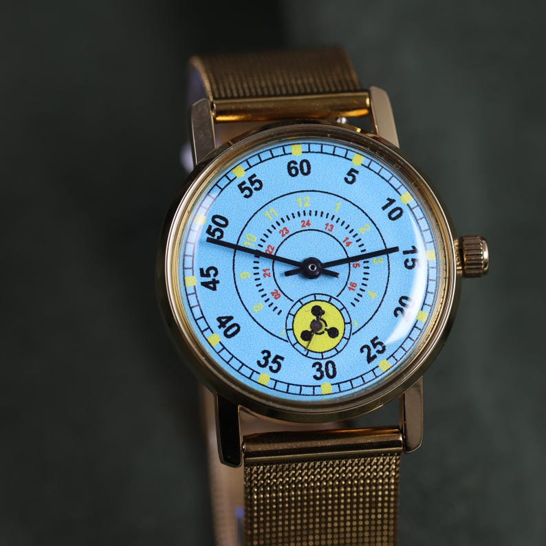 May include: A gold-tone wristwatch with a light blue dial featuring black numerals from 5 to 60, a smaller inner ring with yellow numerals from 8 to 12 and red numerals from 13 to 24, black hands, and a circular yellow icon with a black symbol.