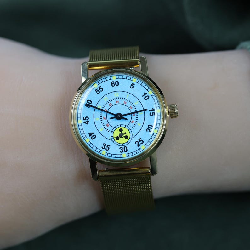 May include: A gold-tone wristwatch with a light blue dial featuring black numerals from 5 to 60, a smaller inner 24-hour scale, and a yellow circular icon with a black three-lobed symbol at the bottom. The watch has a gold mesh metal strap.