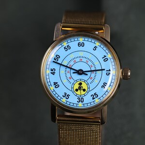 May include: A gold-tone wristwatch with a light blue dial featuring black numerals from 5 to 60, a secondary inner ring with yellow numerals from 1 to 12 and red numerals from 13 to 24, and a small yellow sub-dial with a black three-lobed symbol.