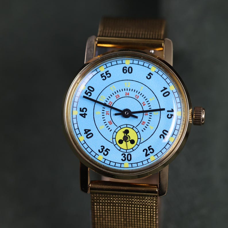 May include: A gold-tone wristwatch with a light blue dial featuring black numerals from 5 to 60, a secondary inner ring with yellow numerals from 1 to 12 and red numerals from 13 to 24, and a small yellow sub-dial with a black three-lobed symbol.