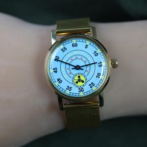 May include: A gold-tone wristwatch with a light blue dial featuring black numerals from 5 to 60, a smaller inner 24-hour ring, and a yellow circular icon with a black three-lobed symbol at the bottom. The watch has a gold mesh metal strap.