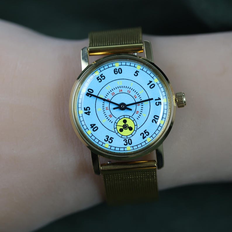 May include: A gold-tone wristwatch with a light blue dial featuring black numerals from 5 to 60, a smaller inner 24-hour ring, and a yellow circular icon with a black three-lobed symbol at the bottom. The watch has a gold mesh metal strap.