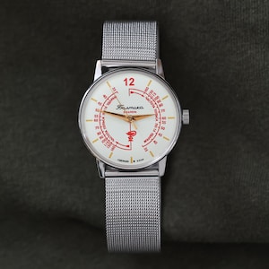 Könnte beinhalten: Eine Vintage-Armbanduhr mit silbernem Mesh-Armband und weißem Zifferblatt. Das Zifferblatt hat rote und goldene Akzente, darunter die Zahl 12 in Rot und kyrillischen Text. Die Uhr ist vor einem dunkelgrünen Hintergrund platziert.