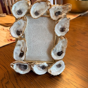 Op de afbeelding: Een rechthoekige fotolijst met een goudkleurige rand versierd met negen oesterschelpen. De randen van elke schelp zijn goudkleurig geschilderd. De inleg van de lijst heeft een wit koraalpatroon en de tekst 4x6 Photo Frame onderaan.