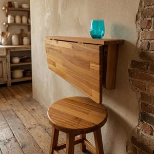 May include: A wall-mounted wooden drop-leaf table with a natural wood grain finish, featuring a hinged rectangular top that folds downwards. A round wooden stool with four legs sits beneath the table, and a teal glass tumbler rests on top.