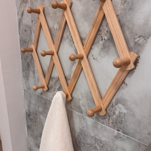 May include: A wooden, expanding wall-mounted coat rack with multiple pegs. The rack is made of light-coloured wood and is mounted on a grey and white tiled wall. A white towel hangs from one of the pegs.