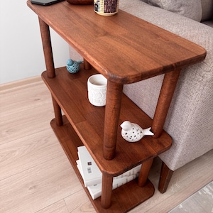 May include: A three-tiered wooden side table with rounded edges and cylindrical legs. The top shelf holds a black smartphone, a small wooden bowl, and a beige mug with coffee bean graphics. The middle shelf holds a white textured cup and a white bird figurine.