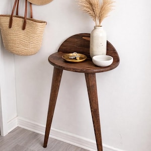 Puede incluir: Una mesa auxiliar de madera marrón oscuro con tres patas anguladas. Una pequeña bandeja dorada con llaves, un cuenco de cerámica blanco y un jarrón moteado con plantas secas se encuentran sobre la mesa. Un sombrero de paja y un bolso de mano tejido cuelgan de la pared.