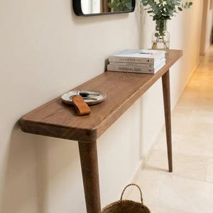 Può includere: Un tavolo console stretto e rettangolare in legno con quattro gambe affusolate. Sulla superficie si trovano un piccolo piatto in ceramica contenente chiavi con un portachiavi in pelle color cuoio, due libri impilati intitolati Kinfolk e Minimalism.