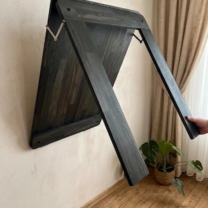 May include: A dark gray, wall-mounted folding table. The table is made of wood and has a rectangular shape. The legs are extended, and the table is mounted on a light-colored wall. The table is designed to fold up against the wall when not in use.