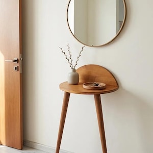 Puede incluir: Una mesa consola de pared de madera en forma de semicírculo con tres patas cónicas, que incluye un pequeño jarrón de cerámica gris claro con ramas de sauce y una bandeja redonda de madera con llaves de metal, bajo un espejo de pared redondo.