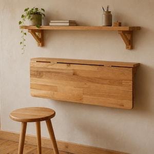 May include: A light-toned wooden wall-mounted drop-leaf table in a folded position, featuring a matching wooden stool with three legs positioned in front. Above the table, a single wooden wall shelf holds a potted plant, books, and a ceramic jar.