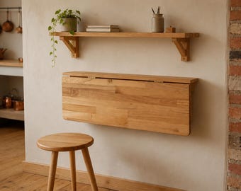 May include: A light-toned wooden wall-mounted drop-leaf table in a folded position, featuring a rectangular surface with visible wood grain. Below the table sits a matching three-legged wooden stool, and above it is a single wooden shelf.
