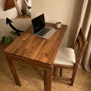 May include: A wooden folding desk and chair set. The desk is rectangular with a dark wood finish, and a laptop and mug are on top. The chair has a light-colored seat and a wooden frame. The desk folds up against the wall.
