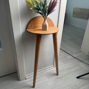 Pode incluir: Uma mesa de consola em madeira semicircular com três pernas cónicas e um pequeno painel traseiro. Um pequeno vaso de cerâmica estriado em tom esbranquiçado assenta sobre o tampo, contendo um arranjo decorativo de ramos artificiais amarelos, verdes e vermelho-escuros.